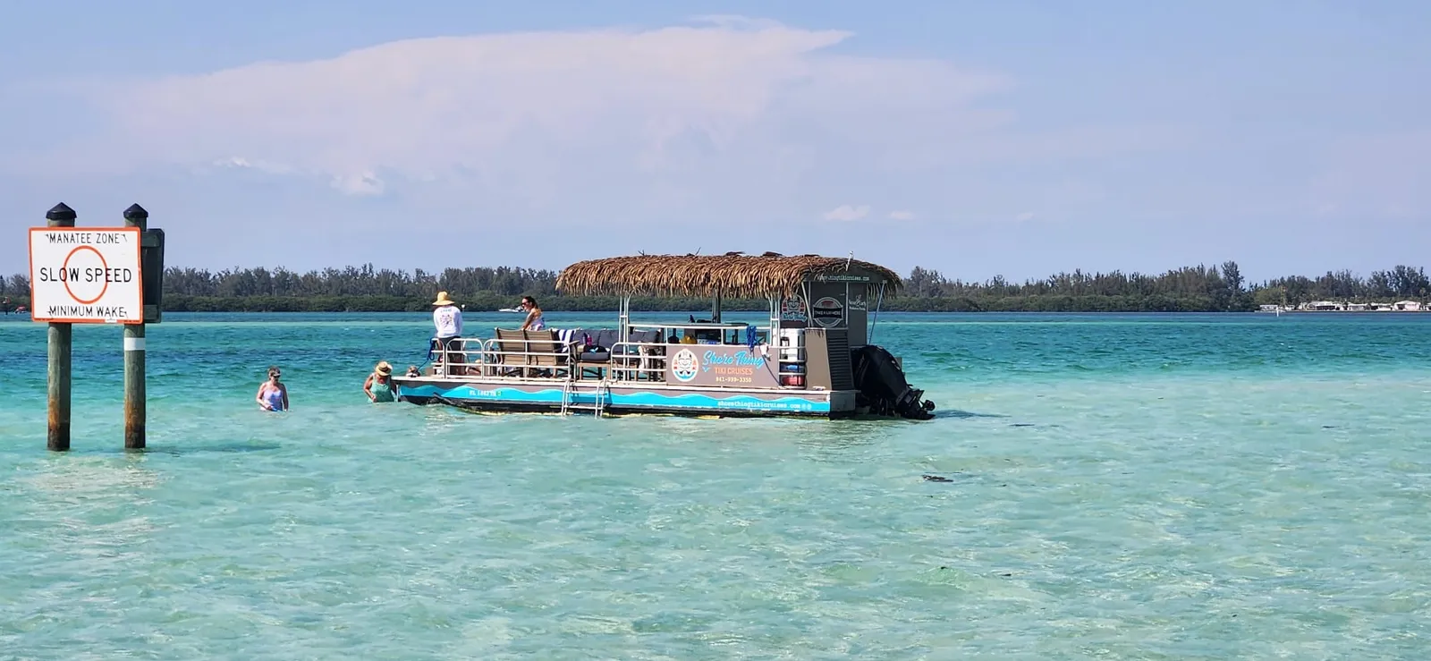 The tiki barge anchored in clear turquoise water beside a manatee zone sign, guests wading alongside