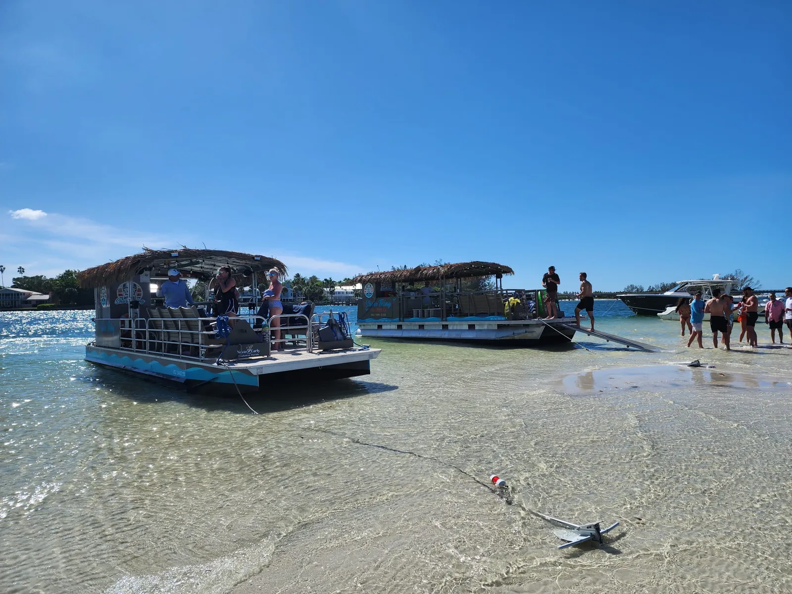 Two tiki barges pulled up on a sandbar under a blue sky, guests stepping off into ankle deep water