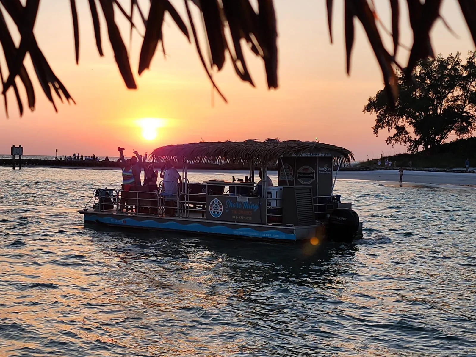 The tiki barge heading out at sunset, guests waving from the bow, seen through palm fronds